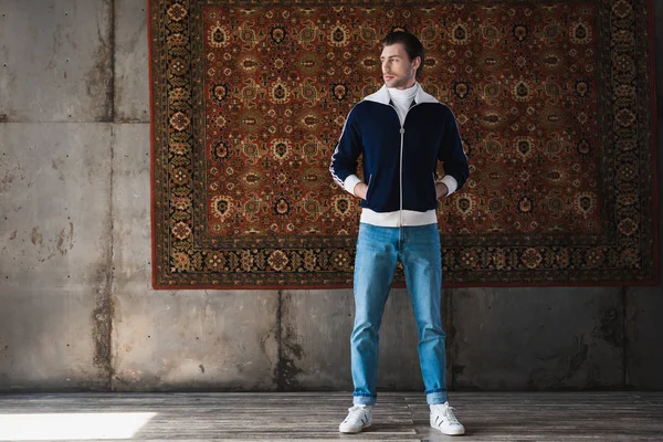 Joven guapo en ropa elegante delante de la alfombra colgando en la pared - foto de stock