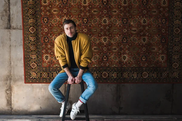 Jeune homme en veste jaune assis sur une chaise devant un tapis accroché au mur — Photo de stock