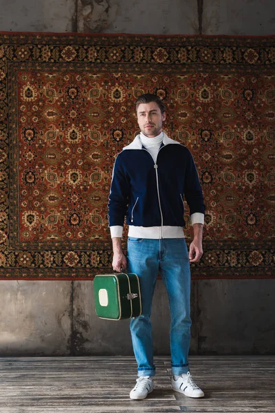 Joven con una pequeña maleta vintage delante de una alfombra colgada en la pared - foto de stock