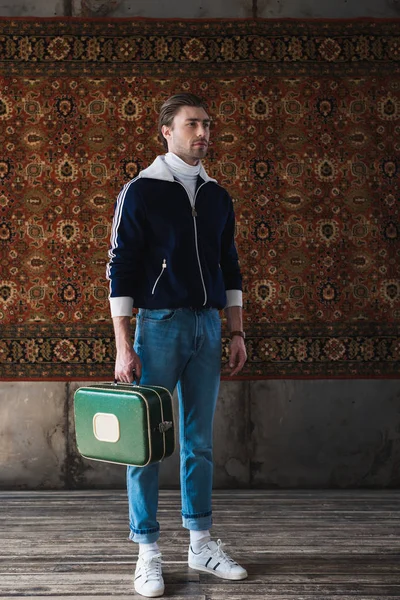 Hombre guapo con la pequeña maleta de la vendimia delante de la alfombra colgando en la pared - foto de stock