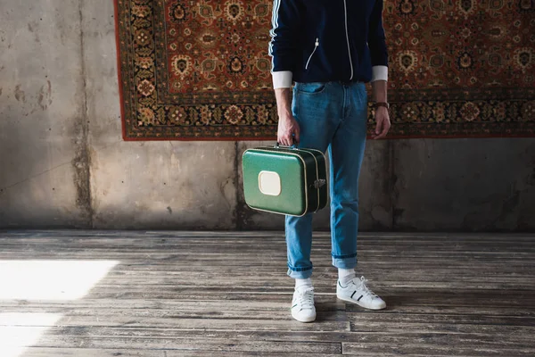 Обрізаний знімок людини з старовинною маленькою валізою перед килимком, що звисає на стіні — стокове фото