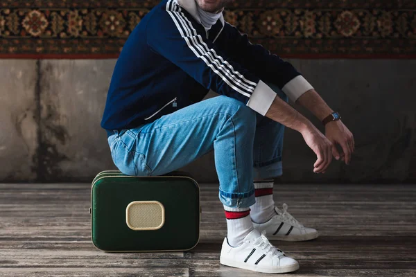 Cropped shot of man sitting on vintage little suitcase in front of rug hanging on wall — Stock Photo