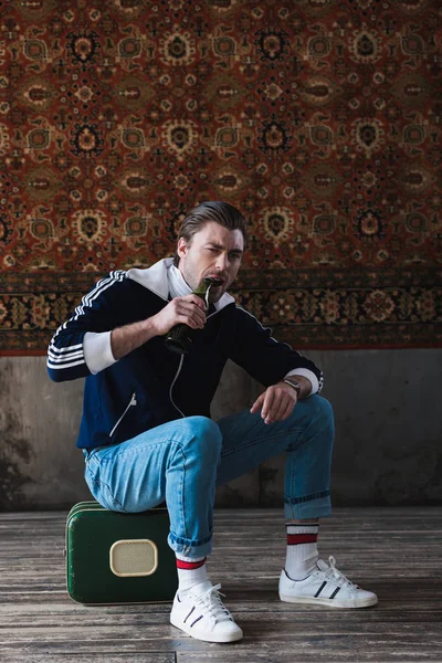 Junger Mann sitzt auf kleinem Vintage-Koffer und versucht, Bier mit Zähnen vor Teppich an der Wand zu öffnen — Stockfoto