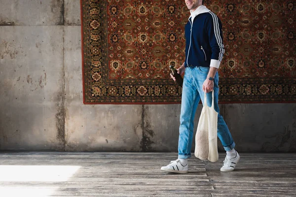 Cropped shot of man in old school clothes with string bag and bottle of beer in front of rug hanging on wall — Stock Photo