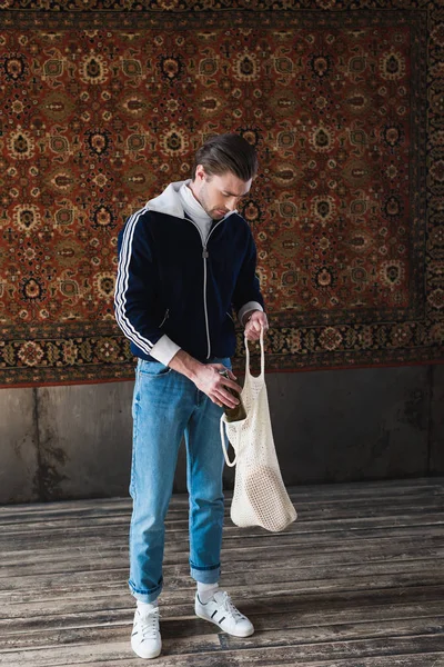 Homme en vêtements old school mettre bouteille de bière dans un sac à ficelle en face du tapis accroché au mur — Photo de stock