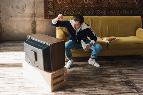 Young man in vintage clothes with bottle of milk punching old tv — Stock Photo