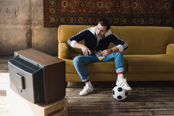 Urlando giovane in abiti vintage con palla guardando il calcio sulla vecchia tv e versando birra in tazza — Foto stock