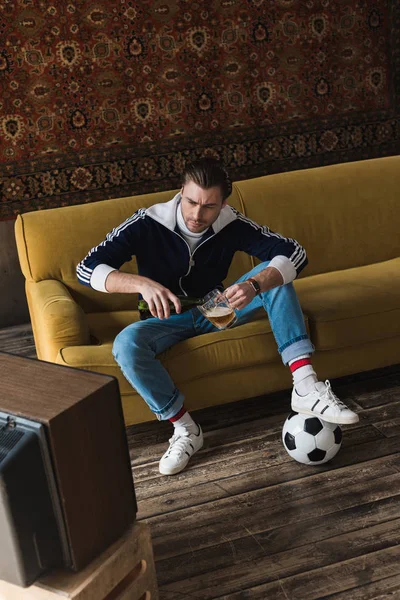 Ernster junger Mann in Vintage-Klamotten mit Ball, der im alten Fernseher Fußball guckt und Bier in Krug gießt — Stockfoto