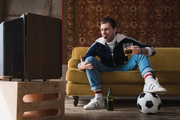 Выразительный молодой человек в винтажной одежде с мячом и кружкой пива смотреть футбол по старому телевизору и кричать — стоковое фото