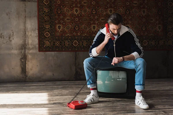 Beau jeune homme en vêtements vintage assis sur le téléviseur rétro et parlant par téléphone et regardant regarder en face du tapis accroché au mur — Photo de stock