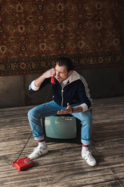 Jeune homme en colère en vêtements vintage assis sur un téléviseur rétro et parlant par téléphone devant un tapis accroché au mur — Photo de stock