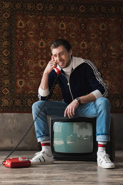 Sorrindo jovem em roupas vintage sentado no aparelho de TV retro e falando por telefone na frente do tapete pendurado na parede — Fotografia de Stock