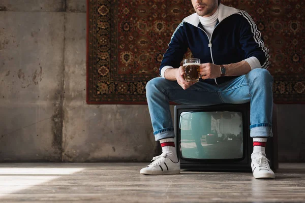 Cropped shot of young man in vintage clothes with mug of beer sitting on retro tv set in front of rug hanging on wall — Stock Photo