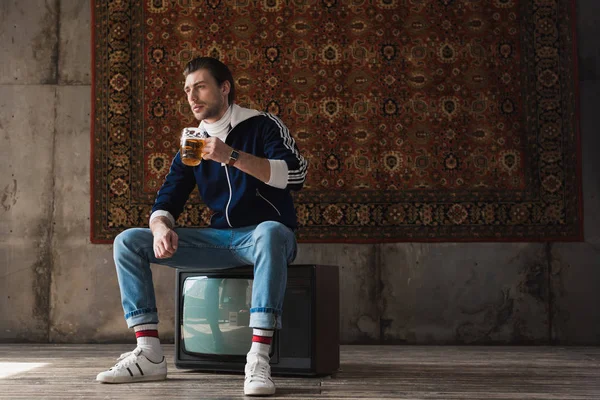 Pensativo joven en ropa vintage con taza de cerveza sentado en retro tv set en frente de la alfombra colgando en la pared - foto de stock