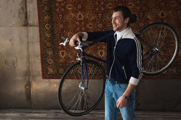 Heureux jeune homme en vêtements vintage portant vélo de route sur l'épaule en face du tapis accroché au mur — Photo de stock