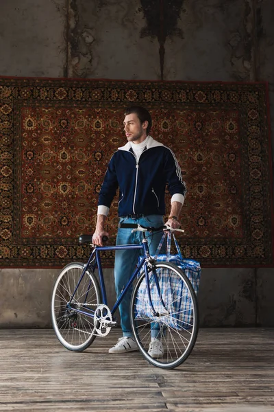 Handsome young man in vintage clothes with road bike and bag in front of rug hanging on wall — Stock Photo