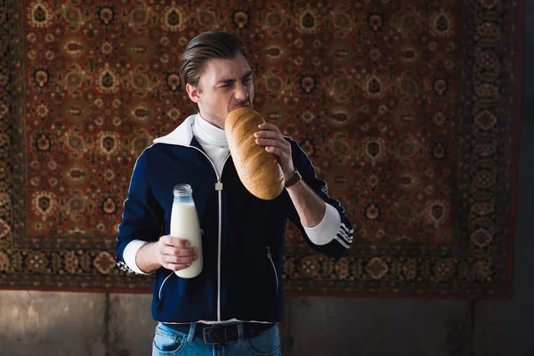 Hungry young man in vintage clothes with bottle of milk biting loaf of bread in front of rug hanging on wall — Stock Photo