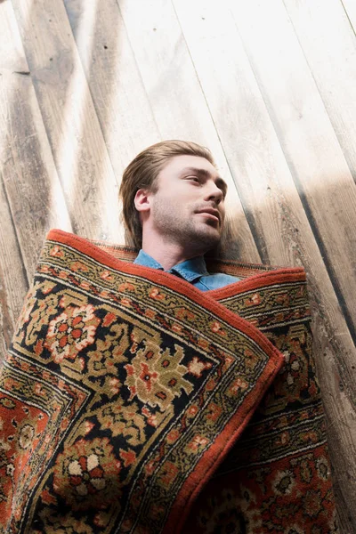 Vista superior de hombre joven cubierto de alfombra acostado en el suelo de madera — Stock Photo