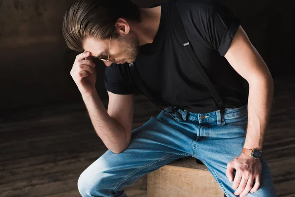 Nachdenklicher junger Mann in schwarzem T-Shirt und Hosenträgern sitzt auf Holzkiste — Stockfoto