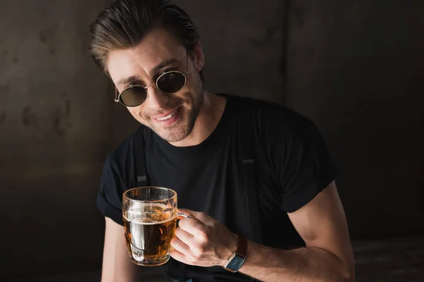 Jovem sorridente em t-shirt preta e óculos de sol segurando caneca de cerveja e olhando para a câmera — Fotografia de Stock
