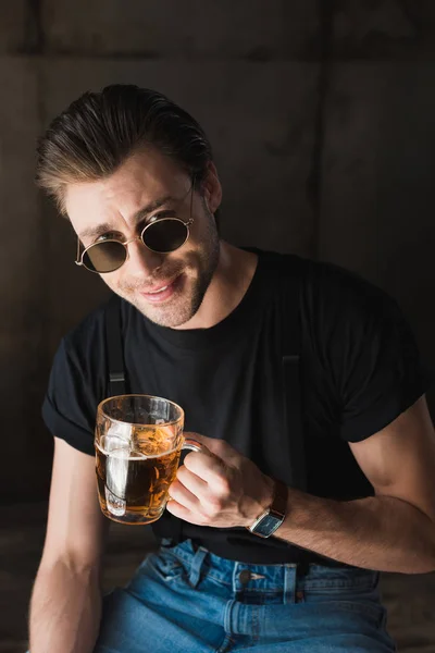 Jovem feliz em t-shirt preta e óculos de sol segurando caneca de cerveja e olhando para a câmera — Fotografia de Stock