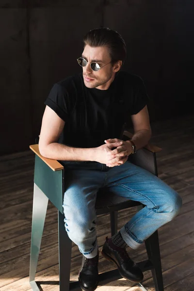 Élégant jeune homme en t-shirt noir et lunettes de soleil assis sur la chaise — Photo de stock