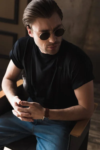 Привабливий молодий чоловік в чорній футболці і сонцезахисних окулярах, сидячи на стільці — стокове фото