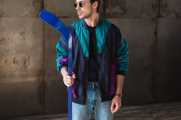 Стильный молодой человек в винтажном ветровщик и солнцезащитные очки с хоккейной клюшкой глядя в сторону — стоковое фото