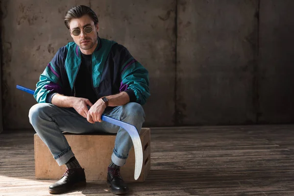 Schöner junger Mann in Vintage-Windjacke mit Hockeyschläger sitzt auf Holzkiste — Stockfoto