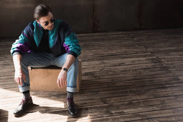 Stylish young man in vintage windcheater sitting on wooden box in loft room — Stock Photo