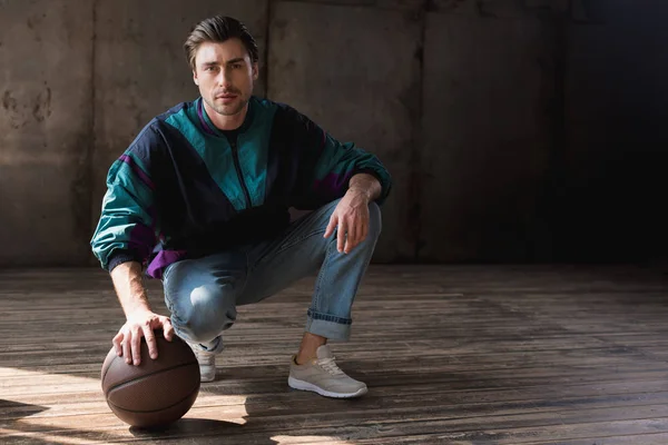 Attractive young man in vintage windcheater with basketball ball sitting squats on wooden floor — Stock Photo