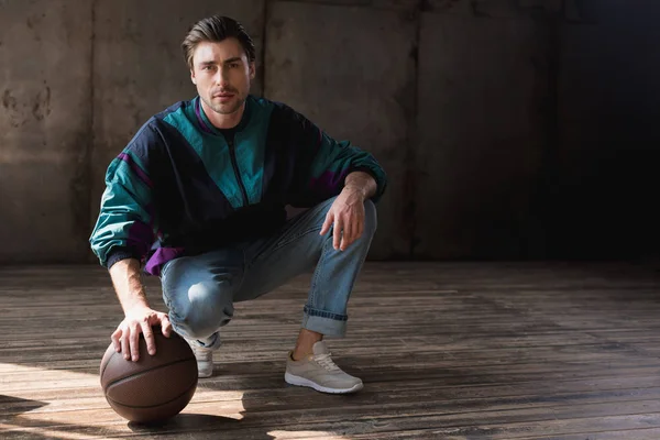 Atractivo joven en vintage windcheater con pelota de baloncesto sentados sentados sentados en piso de madera - foto de stock