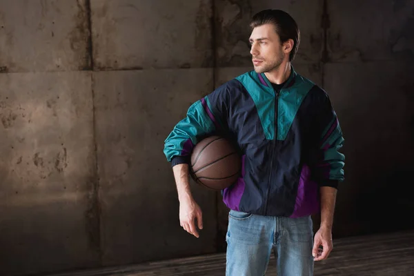 Стильный молодой человек в винтажном ветровщике с баскетбольным мячом, смотрящим в сторону — стоковое фото