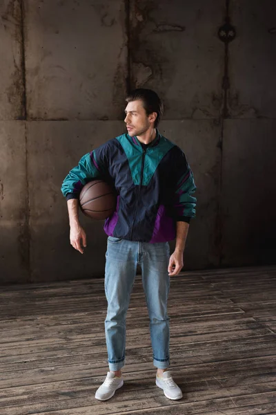 Atractivo joven en vintage windcheater con pelota de baloncesto mirando hacia otro lado - foto de stock