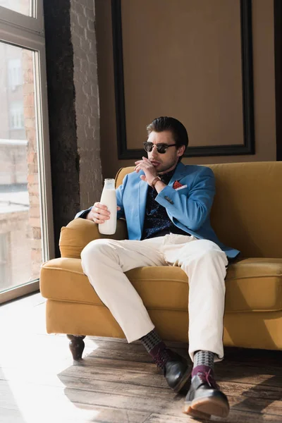 Привабливий молодий чоловік в стильному костюмі сидить на дивані з пляшкою молока і протирає рот рукою — стокове фото