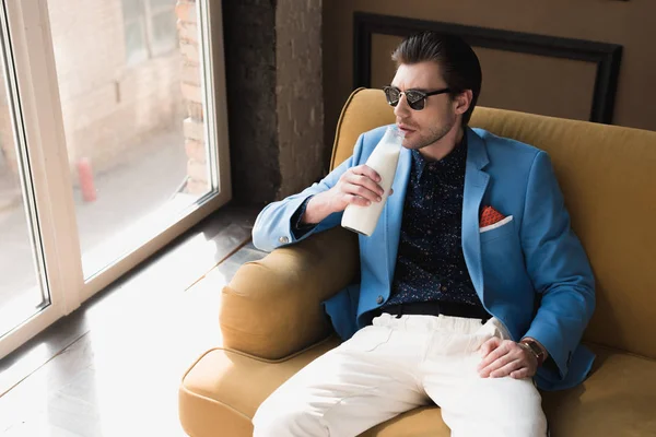 Vista de ángulo alto del joven guapo en traje elegante sentado en el sofá y beber leche de la botella - foto de stock