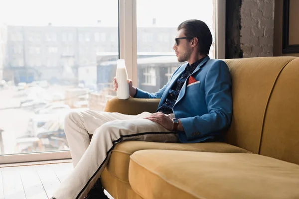 Nachdenklicher junger Mann im eleganten Anzug sitzt mit einer Flasche Milch auf dem Sofa und schaut durch das Fenster — Stockfoto
