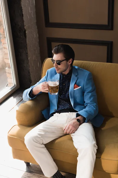 Vista de ángulo alto de joven guapo en traje elegante sentado en el sofá con taza de cerveza - foto de stock