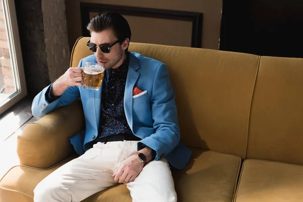 Attractive young man in stylish suit sitting on couch and drinking beer — Stock Photo