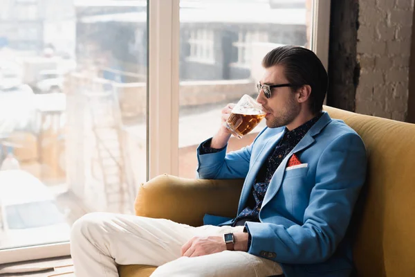 Bello giovane in abito elegante seduto sul divano e bere birra — Foto stock