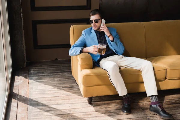 Stylish young man with mug of beer talking by phone while sitting on couch — Stock Photo