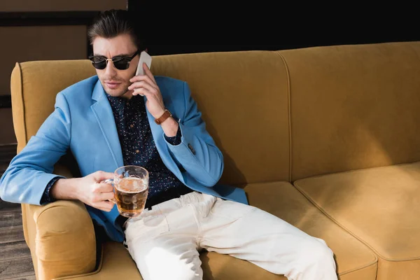 Young man with mug of beer talking by phone while sitting on couch — Stock Photo