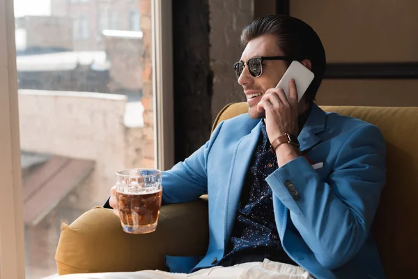Красивий молодий чоловік з чашкою пива розмовляє по телефону, сидячи на дивані — стокове фото