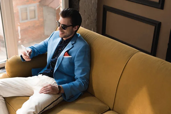 Blick aus der Vogelperspektive auf einen lächelnden jungen Mann im eleganten Anzug mit Smartphone, während er auf der Couch sitzt — Stockfoto