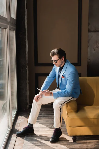 Schöner junger Mann im schicken Anzug mit Smartphone auf der Couch sitzend — Stockfoto