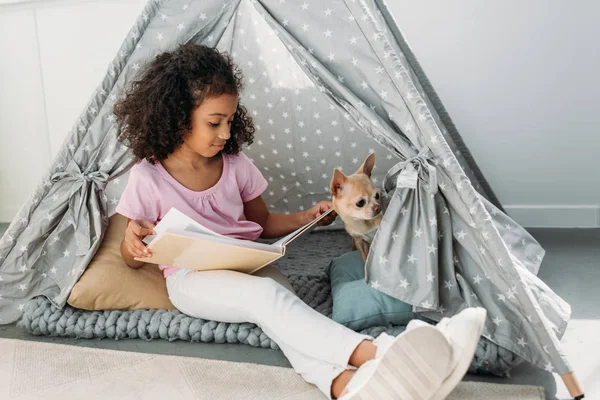 Little african american kid reading book with chihuahua dog near by in teepee at home — Stock Photo