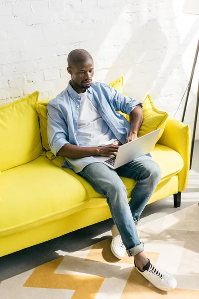 Uomo afro-americano che utilizza il computer portatile sul divano di casa — Foto stock