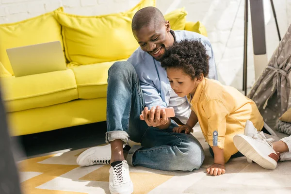 Улыбающиеся африканские американец и маленький сын, используя смартфон вместе дома — стоковое фото