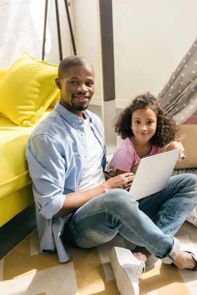 Père afro-américain avec ordinateur portable et fille avec chien assis sur le sol ensemble à la maison — Photo de stock