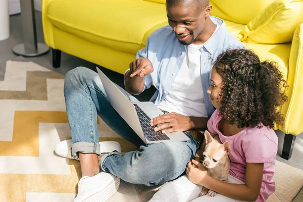 Афроамериканський батько і маленька дочка з собакою використовують ноутбук разом вдома — стокове фото